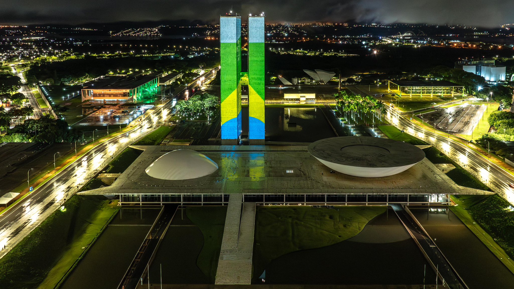 Foto noturna do Eixo Monumental, com foco no Congresso Nacional. Há uma projeção nos prédios do Congresso Nacional com a imagem da bandeira do Brasil.